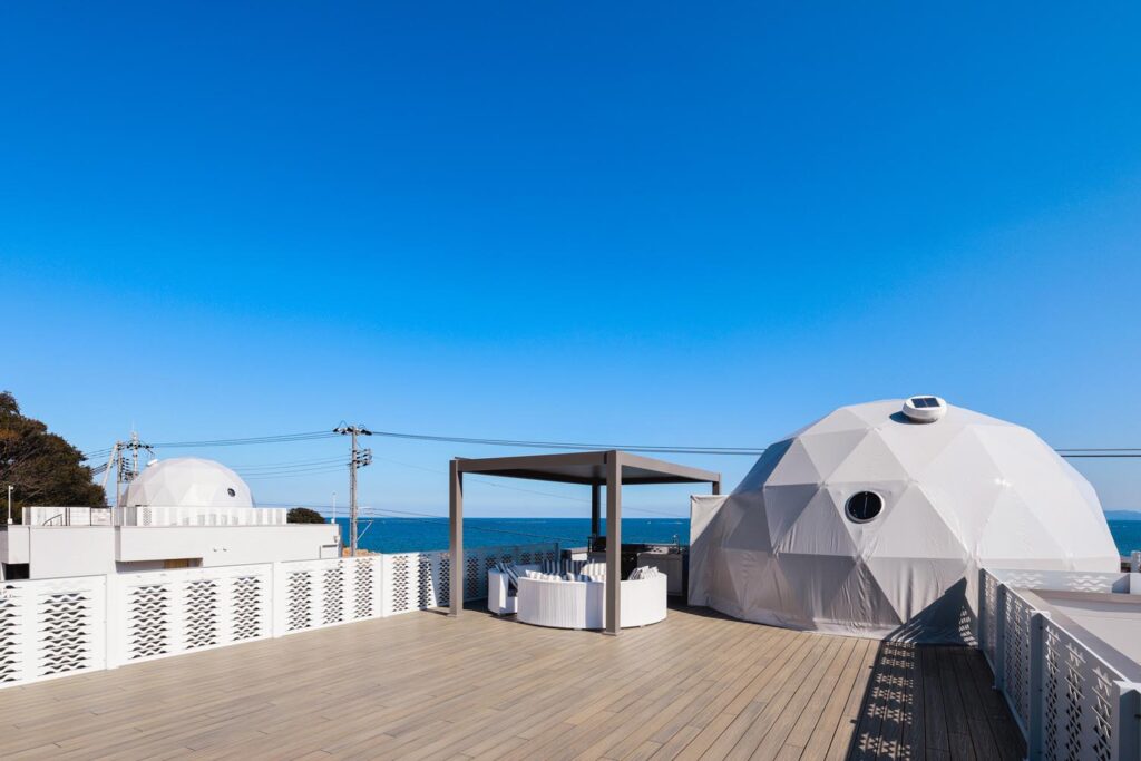 海を望むテラスの屋外リビングスペース。白いラタンのソファセットとグリルキッチン、ドーム型のテントが見える。