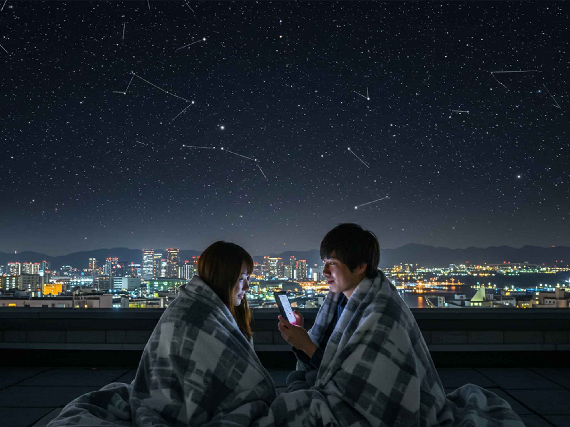 淡路島の街夜景と星空を背景に、カップルが屋上でブランケットにくるまって星空観賞。冬の服装や観賞準備の参考になるシーン。