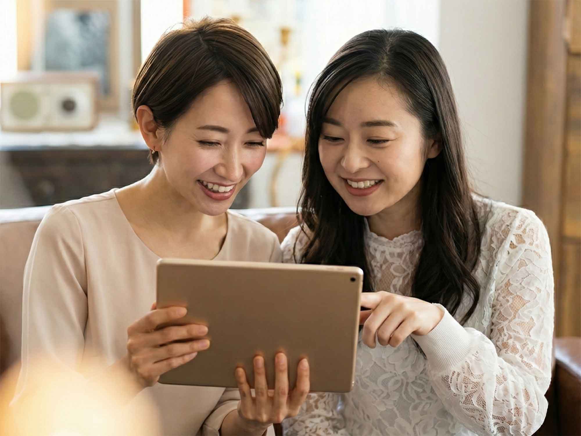 室内で二人の女性が並んで座り、タブレット端末を手に持って画面を見ている。背景には家具や棚があり、家庭内の落ち着いた空間で情報を確認している様子が写っている。