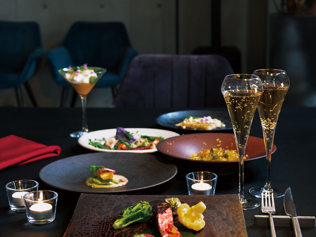 落ち着いた雰囲気の黒いテーブルの上に、コース料理とシャンパンが並べられている。手前にはキャンドルとカトラリーがあり、奥には青い椅子がいくつか見える。