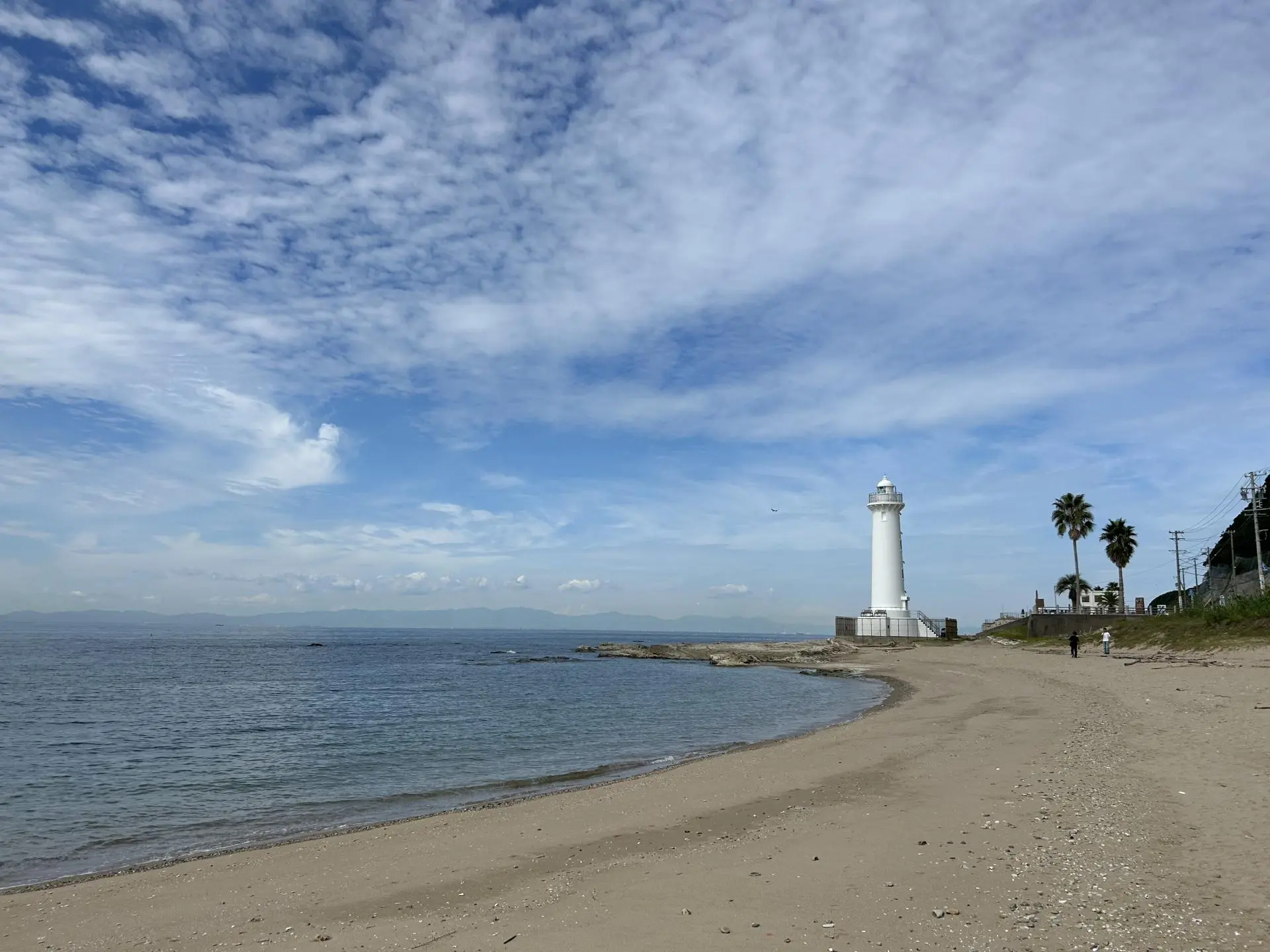 white-lighthouse-on-sandy-beach - CORAL RESORT コーラルリゾート 砂浜の海岸に立つ白い灯台と、遠くに見える山々。青い空には白い雲が広がり、海は穏やかだ。ヤシの木と二人の人物も見える。